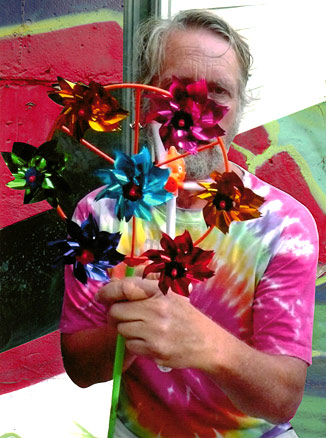 Dan with one eye visible, holding large multi-colored pin wheel, multi-colored graffiti behind him.
