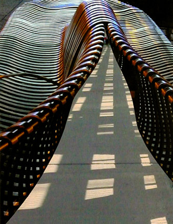 Two metal benches converge, their forms made of struts like zipper teeth, open onto a path with shadow stripes.